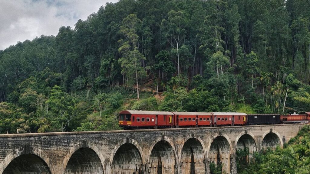 A red train crossing the Nine Arches Bridge in Ella, Sri Lanka, surrounded by lush green bushes and scenic hill country landscape.